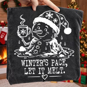 May include: A dark gray t-shirt featuring a white graphic of a melting snowman wearing a winter hat, holding a steaming mug with a snowflake design. Below the snowman, the text reads: WINTER'S PACE, LET IT MELT. A small heart icon is centered below the text.