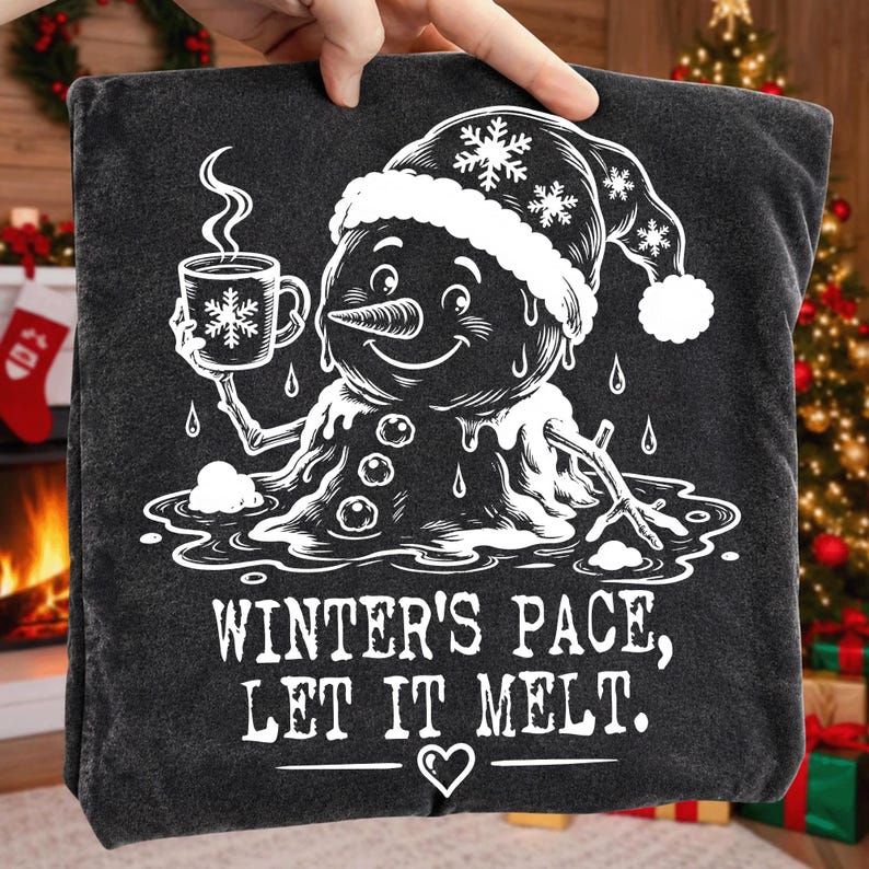 May include: A dark gray t-shirt featuring a white graphic of a melting snowman wearing a winter hat, holding a steaming mug with a snowflake design. Below the snowman, the text reads: WINTER'S PACE, LET IT MELT. A small heart icon is centered below the text.