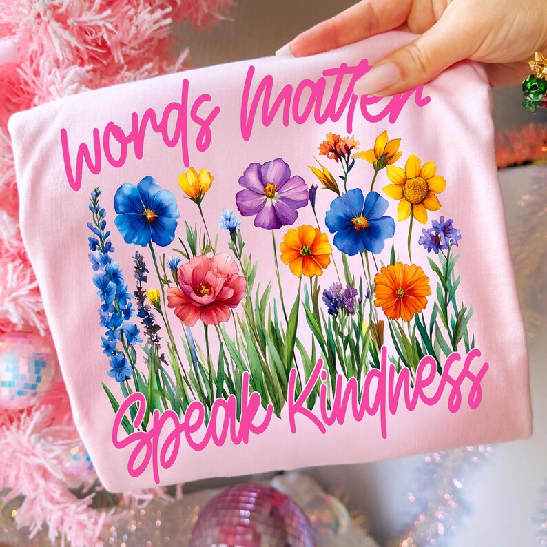 Puede incluir: Una camiseta rosa claro con las palabras "Words Matter" y "Speak Kindness" en escritura rosa. La camiseta presenta un dise&ntilde;o floral colorido con flores azules, amarillas, naranjas y moradas.