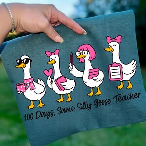 K&ouml;nnte beinhalten: T&uuml;rkisfarbenes T-Shirt mit einem Design von vier Cartoon-G&auml;nsen. Jede Gans hat ein anderes Accessoire, wie eine Sonnenbrille, eine rosa Schleife oder einen Klemmbrett. Der Text "100 Days. Same Silly Goose Teacher" ist darunter gedruckt.