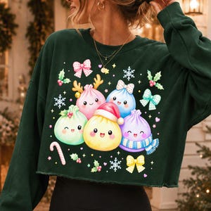 May include: A dark green long-sleeve cropped sweatshirt featuring a graphic print of five colourful, smiling dumpling characters. The dumplings are decorated with festive elements including a Santa hat, reindeer antlers, a scarf, and bows, surrounded by snowflakes and holly.
