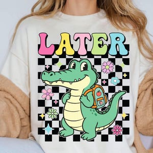 K&ouml;nnte beinhalten: Cremefarbenes Sweatshirt mit einer bunten Grafik. Das Design zeigt das Wort "LATER" in Buchstabenblasen &uuml;ber einem Cartoon-Alligator mit Rucksack. Der Hintergrund ist ein schwarz-wei&szlig;es Schachbrettmuster mit floralen und sternf&ouml;rmigen Akzenten.