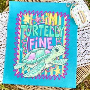 May include: A turquoise tote bag with a cartoon turtle illustration and the phrase "I'M TURTELLY FINE" in colorful text. The turtle is light green and pink, with a smiling face. The background has vertical stripes and stars.