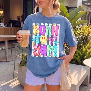 May include: A blue t-shirt with the text "BORN TO YAP!" in colourful, playful letters. The design includes a smiley face, stars, and a bow. The shirt is paired with striped shorts.