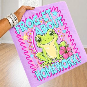 K&ouml;nnte beinhalten: Eine hellviolette Decke mit dem Text "FROG-ET ABOUT HOMEWORK!" in T&uuml;rkis und Pink. In der Mitte befindet sich eine Cartoon-Frosch mit einer rosa Blume. Die Decke wird von einer Person gehalten.