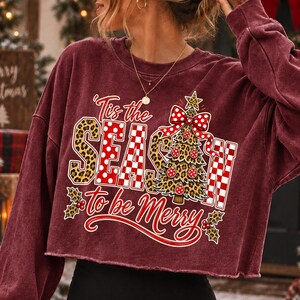 Op de afbeelding: Een bordeauxrood, langmouwig, kort sweatshirt met een grafische print met de tekst 'Tis the SEASON to be Merry'. Het ontwerp bevat een luipaardprint, rood-witte stippen, een kerstboom en hulstbladaccenten.
