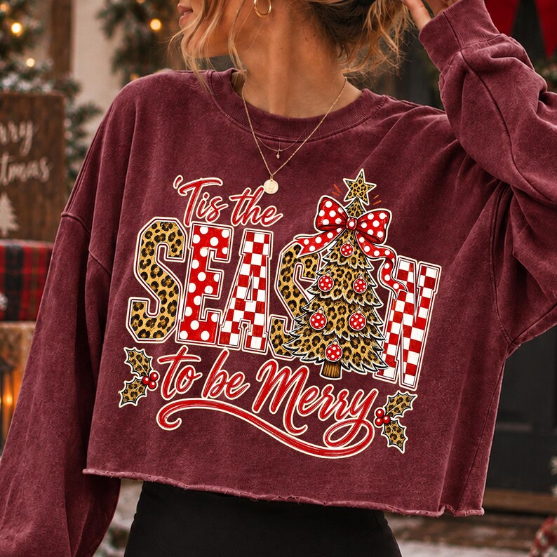 Op de afbeelding: Een bordeauxrood, langmouwig, kort sweatshirt met een grafische print met de tekst 'Tis the SEASON to be Merry'. Het ontwerp bevat een luipaardprint, rood-witte stippen, een kerstboom en hulstbladaccenten.