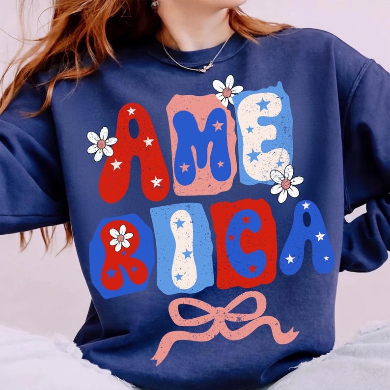 May include: Navy blue sweatshirt featuring the word "AMERICA" in red, white, and blue bubble letters. The letters are decorated with stars and flowers, and a pink bow is at the bottom. A casual, patriotic design.