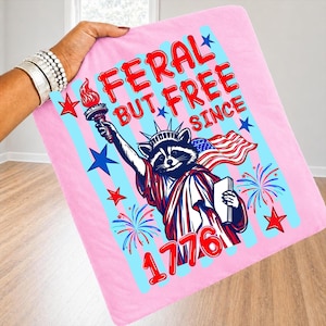 May include: Pink blanket featuring a raccoon dressed as the Statue of Liberty, holding a torch and the American flag. Text reads "FERAL BUT FREE SINCE 1776" in red and blue. Stars and fireworks complete the patriotic design.