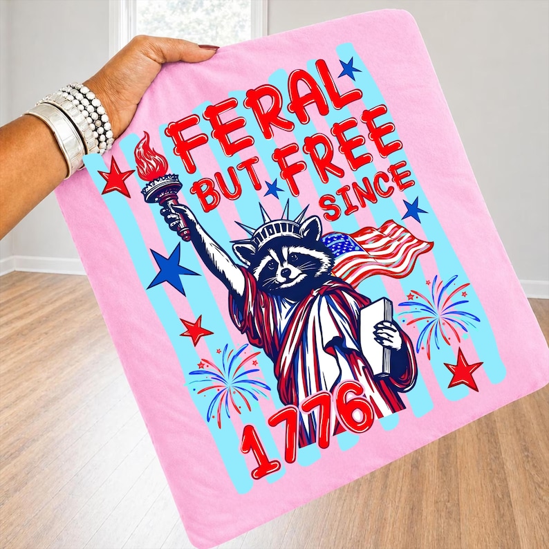 May include: Pink blanket featuring a raccoon dressed as the Statue of Liberty, holding a torch and the American flag. Text reads "FERAL BUT FREE SINCE 1776" in red and blue. Stars and fireworks complete the patriotic design.