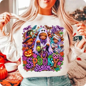 Może przedstawiać: Biała bluza z długim rękawem z kolorową grafiką przedstawiającą trzy krowy rasy Highland w kostiumach na Halloween, w tym czarownicę, ducha i mumię, z napisem "SPOOKY SEASON" wykonanym żywymi, wielokolorowymi literami poniżej.