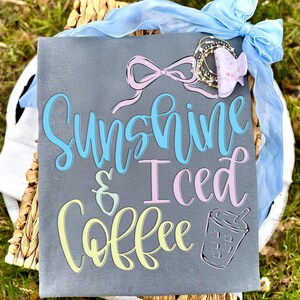 Op de afbeelding: Een grijze canvas met de tekst "Sunshine & Iced Coffee" in blauwe, gele en roze letters. Een tekening van een ijskoffiebeker staat rechts. Een lichtblauw lint en een decoratieve haarclip staan erboven.