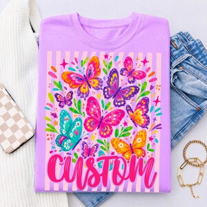May include: A light purple t-shirt featuring a graphic print of multiple colourful butterflies in shades of pink, orange, yellow, teal, and purple, accented with green leaves and small stars, with the word CUSTOM written in a bold pink script font.