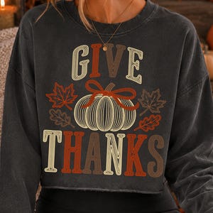 Puede incluir: Una sudadera de manga larga gris carb&oacute;n con un estampado gr&aacute;fico que incluye el texto GIVE THANKS en una fuente retro. El dise&ntilde;o presenta una calabaza central con l&iacute;neas blancas, un lazo rojo y hojas de arce y roble en tonos naranja y rojo.