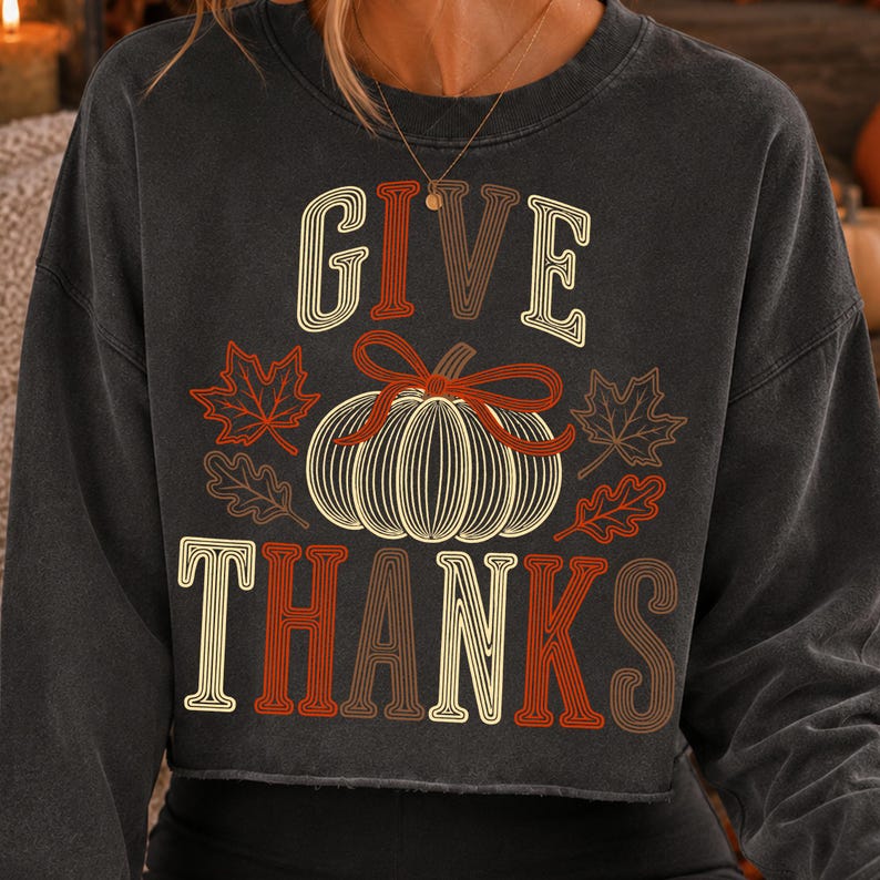 Puede incluir: Una sudadera de manga larga gris carb&oacute;n con un estampado gr&aacute;fico que incluye el texto GIVE THANKS en una fuente retro. El dise&ntilde;o presenta una calabaza central con l&iacute;neas blancas, un lazo rojo y hojas de arce y roble en tonos naranja y rojo.