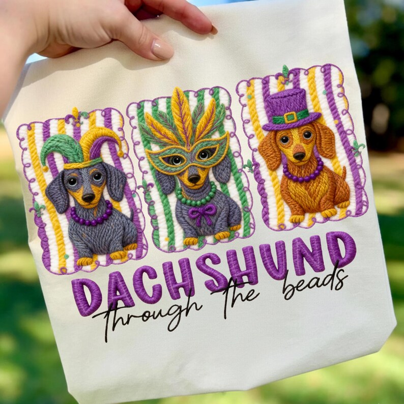 Puede incluir: Bolso tote color crema con tres perros salchicha bordados con disfraces de Mardi Gras. Cada perro lleva un accesorio festivo diferente: un gorro de buf&oacute;n, una m&aacute;scara de plumas y un sombrero de copa. El texto "DACHSHUND Through The beads" est&aacute; bordado debajo.