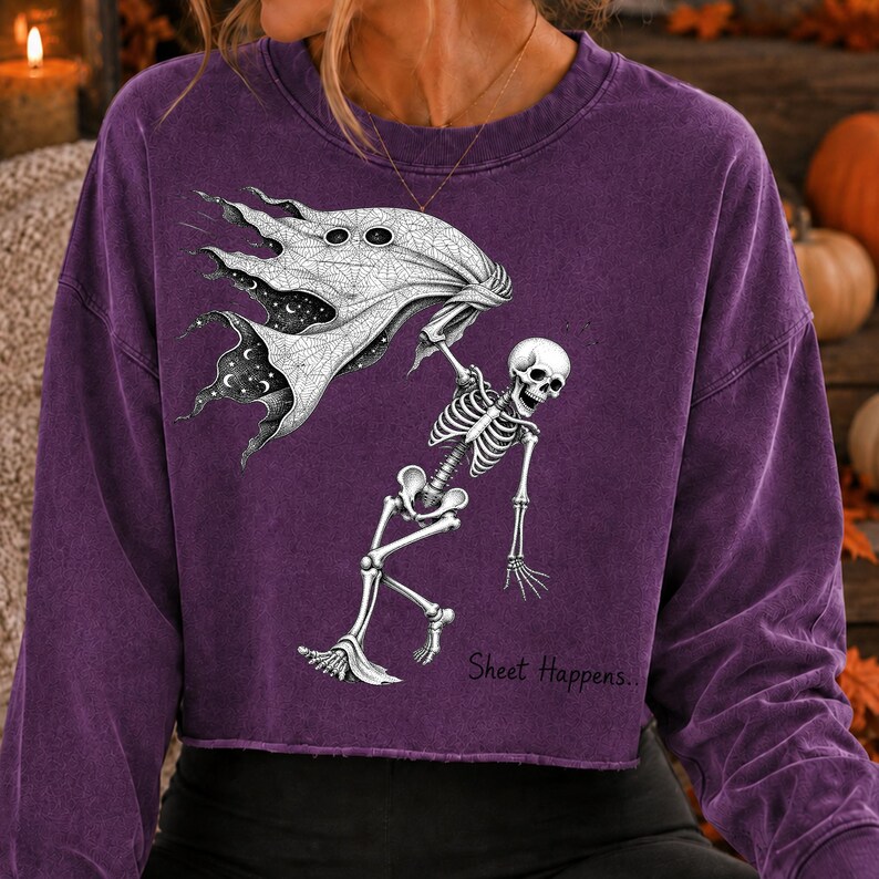 K&ouml;nnte beinhalten: Ein violettes Langarm-Sweatshirt mit Rundhalsausschnitt und offenem Saum. Die Grafik zeigt ein wei&szlig;es Skelett, das ein flie&szlig;endes Laken mit Sternenhimmel-Muster h&auml;lt. Der Text "Sheet Happens.." ist in Schwarz unter der Skelettfigur aufgedruckt.