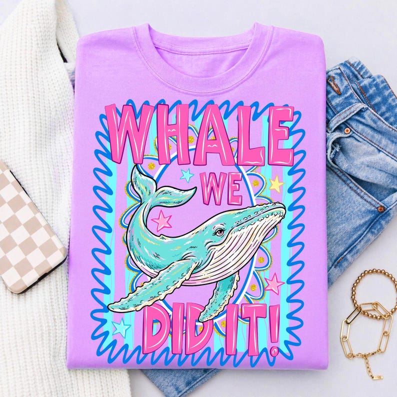 K&ouml;nnte beinhalten: Ein lilafarbenes T-Shirt mit einer t&uuml;rkisfarbenen Walillustration und dem pinkfarbenen Text "WHALE WE DID IT!". Das Design umfasst Sterne und einen blauen Wellenrand. Das Shirt wird mit einer blauen Jeans pr&auml;sentiert, was einen l&auml;ssigen und farbenfrohen Look erzeugt.