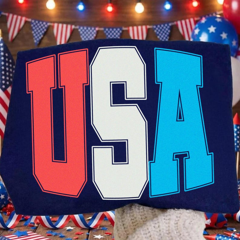 May include: A navy blue item with the word "USA" in large, bold letters. The letters are outlined in white, with the "U" in red, the "S" in white, and the "A" in blue. The background includes American flags and festive decorations.