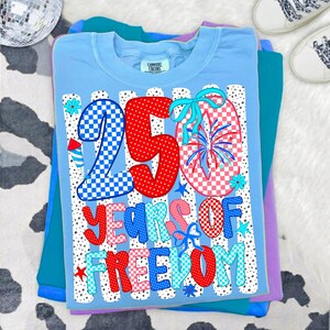 K&ouml;nnte beinhalten: Ein hellblaues T-Shirt mit einem Aufdruck, der den Text 250 YEARS OF FREEDOM zeigt. Das Design umfasst rot-wei&szlig;-blaue Karomuster, Punkte, Feuerwerksdarstellungen und kleine blaue Schleifen auf einem Hintergrund aus vertikalen Streifen.