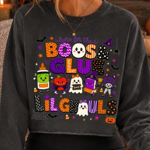 K&ouml;nnte beinhalten: Ein anthrazitfarbenes Langarm-Sweatshirt mit einer bunten Halloween-Grafik und dem Text "here for the BOOS & GLUE with my LIL GHOULS". Das Design zeigt Zeichentrickfiguren wie ein Gespenst, eine Katze und eine Klebeflasche sowie Flederm&auml;use und Sterne.