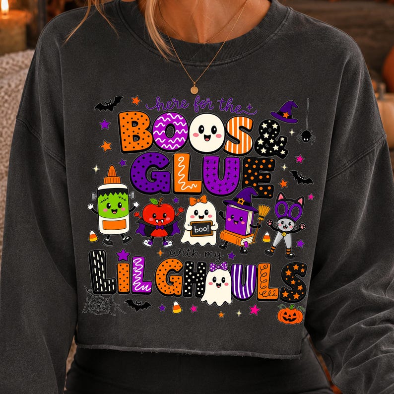 K&ouml;nnte beinhalten: Ein anthrazitfarbenes Langarm-Sweatshirt mit einer bunten Halloween-Grafik und dem Text "here for the BOOS & GLUE with my LIL GHOULS". Das Design zeigt Zeichentrickfiguren wie ein Gespenst, eine Katze und eine Klebeflasche sowie Flederm&auml;use und Sterne.