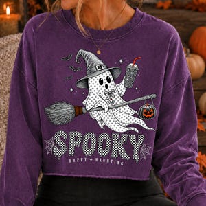 May include: A purple long-sleeve sweatshirt featuring a graphic of a ghost wearing a witch hat, holding a drink and a broomstick with a jack-o'-lantern. The text reads SPOOKY HAPPY HAUNTING, accompanied by small bats, stars, and spider web designs.