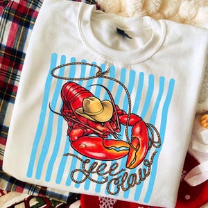 Op de afbeelding: Wit sweatshirt met een rode kreeft met een gele cowboyhoed, een touwlasso en blauwe verticale strepen. De tekst "Yee Claw" is geschreven in een touwachtig lettertype. Een leuk, casual ontwerp.