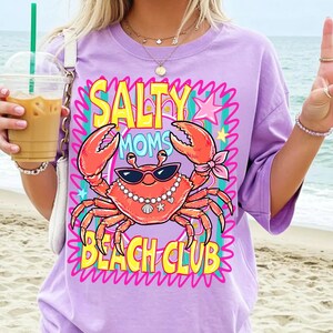 May include: A lilac t-shirt featuring a cartoon crab wearing sunglasses and a pearl necklace. The text "SALTY MOMS BEACH CLUB" is in yellow and pink. The crab is orange with pink details, evoking a beach theme.