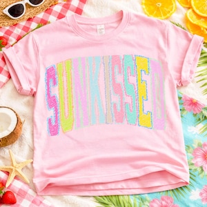 May include: A pale pink t-shirt featuring the word "SUNKISSED" in a stitched, rainbow-coloured design. The letters are in shades of blue, yellow, green, and pink. The sleeves are rolled up. The shirt is on a beach-themed background.