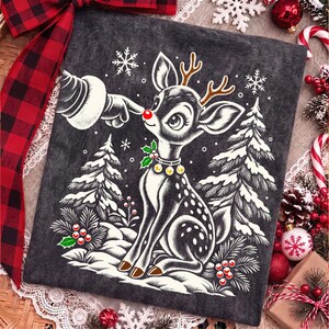 Puede incluir: Una camiseta gris oscuro con una ilustraci&oacute;n gr&aacute;fica de un cervatillo con una nariz roja brillante siendo tocado por un dedo enguantado. El dise&ntilde;o incluye astas marrones, un collar con cascabeles, copos de nieve blancos y pinos nevados.