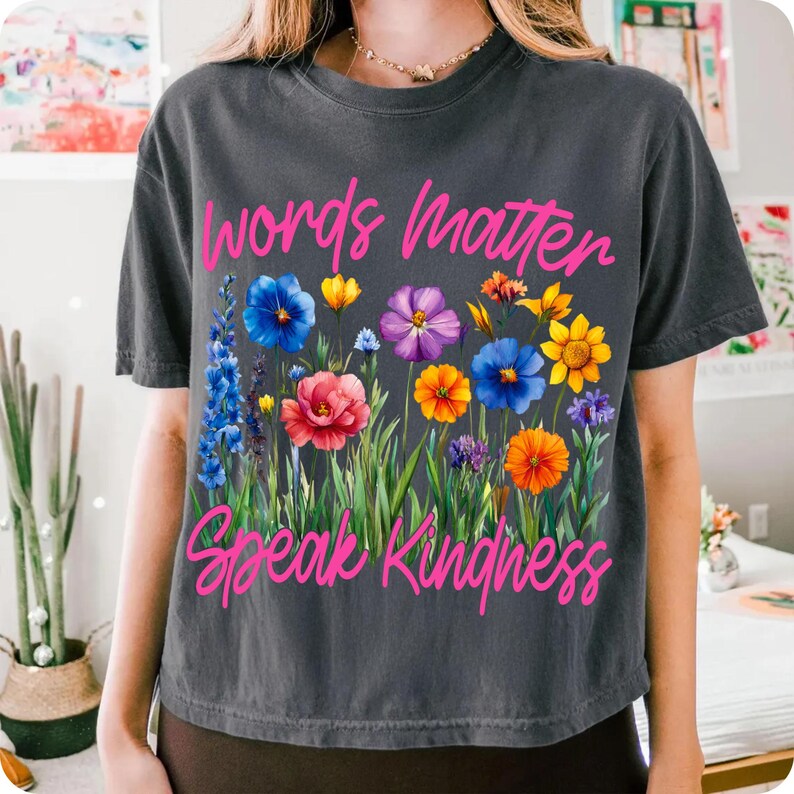 Puede incluir: Camiseta gris oscuro con un dise&ntilde;o floral y el texto rosa "Words Matter Speak Kindness". El dise&ntilde;o floral incluye flores azules, amarillas, moradas, naranjas y rojas con tallos verdes.