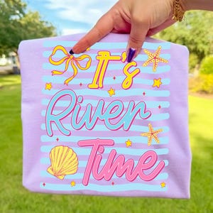 May include: A light purple t-shirt with the text "It's River Time" in yellow, blue, and pink. The design includes a yellow bow, starfish, a seashell, and stars. The shirt has light blue horizontal stripes.