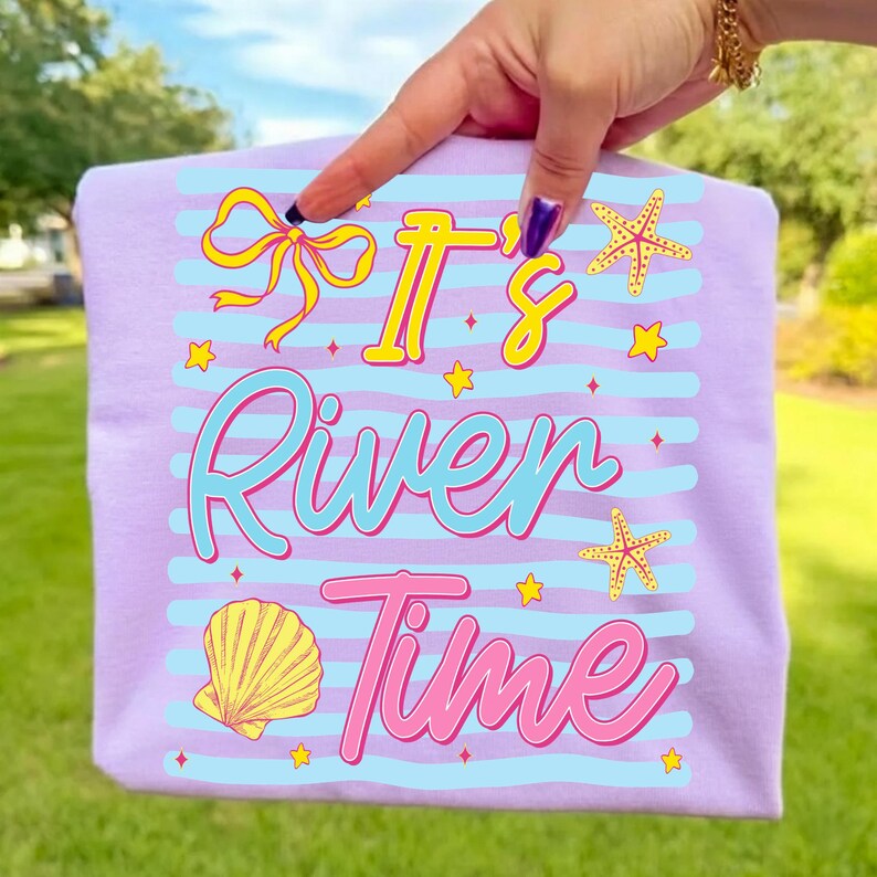 May include: A light purple t-shirt with the text "It's River Time" in yellow, blue, and pink. The design includes a yellow bow, starfish, a seashell, and stars. The shirt has light blue horizontal stripes.