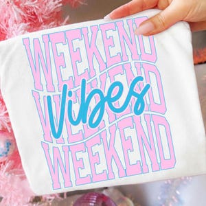 K&ouml;nnte beinhalten: Wei&szlig;es T-Shirt mit den Worten "WEEKEND VIBES WEEKEND" in einem wellenf&ouml;rmigen Design. Der Text ist in hellblauer Schrift und rosa umrandet. Das T-Shirt wird von einer Hand gehalten.