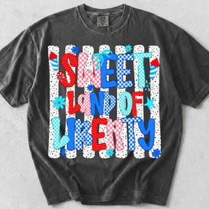 May include: A charcoal grey short-sleeve t-shirt featuring a graphic print with the text SWEET LAND OF LIBERTY in red, white, and blue checkered and polka dot patterns. The design includes firework and star motifs over a vertical striped background.