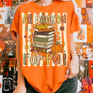 Op de afbeelding: Een oranje t-shirt met een afbeelding van een stapel van vijf boeken en een pompoen. De tekst luidt All Booked For Fall. Het ontwerp bevat herfstbladeren en een gestreept achtergrondpatroon. Het t-shirt heeft korte mouwen en een ronde hals.