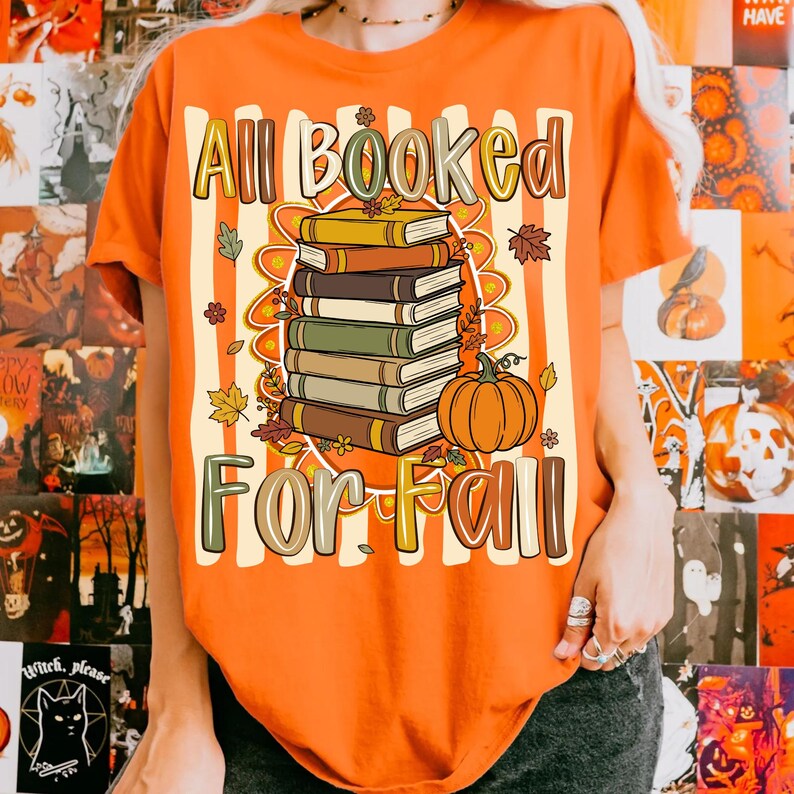 Op de afbeelding: Een oranje t-shirt met een afbeelding van een stapel van vijf boeken en een pompoen. De tekst luidt All Booked For Fall. Het ontwerp bevat herfstbladeren en een gestreept achtergrondpatroon. Het t-shirt heeft korte mouwen en een ronde hals.