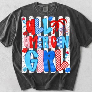 Pode incluir: Camiseta cinza escuro com o texto "ALL AMERICAN GIRL" em letras vermelhas, azuis e rosa. As letras t&ecirc;m padr&otilde;es xadrez e listrados. O design inclui fogos de artif&iacute;cio, flores e um la&ccedil;o vermelho.