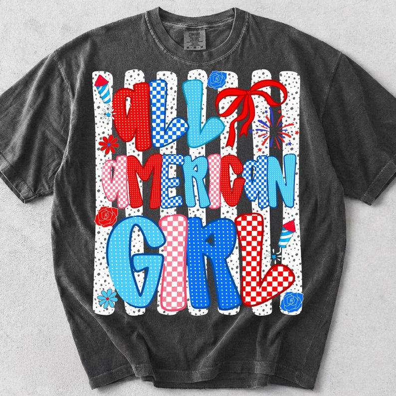 Pode incluir: Camiseta cinza escuro com o texto "ALL AMERICAN GIRL" em letras vermelhas, azuis e rosa. As letras t&ecirc;m padr&otilde;es xadrez e listrados. O design inclui fogos de artif&iacute;cio, flores e um la&ccedil;o vermelho.