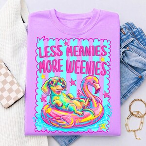 May include: A light purple t-shirt featuring a graphic of a dachshund wearing heart-shaped sunglasses while floating on a pink flamingo pool float. The text "LESS MEANIES MORE WEENIES" is printed in pink above the dog, surrounded by a blue striped border.