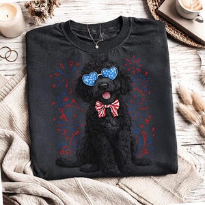 Pu&ograve; includere: Maglietta nera con un cane nero con occhiali a forma di cuore e un papillon rosso, bianco e blu. Fuochi d'artificio rossi e blu circondano il cane. &Egrave; visibile una collana.