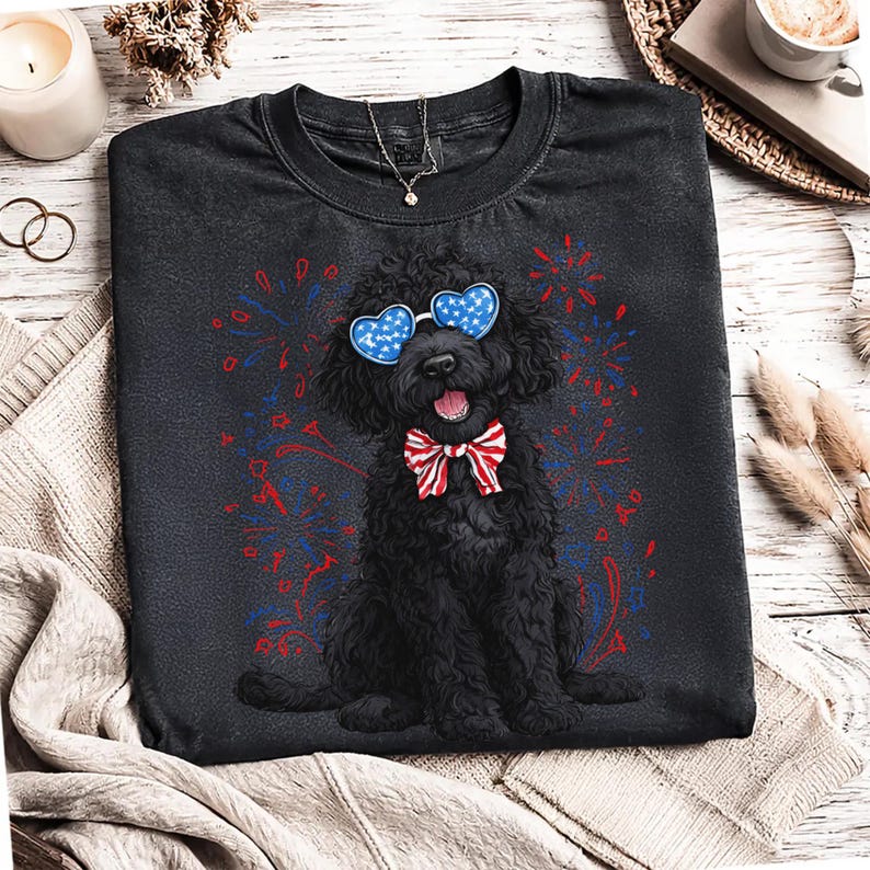 Pu&ograve; includere: Maglietta nera con un cane nero con occhiali a forma di cuore e un papillon rosso, bianco e blu. Fuochi d'artificio rossi e blu circondano il cane. &Egrave; visibile una collana.