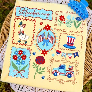 May include: A yellow decorative pillow with patriotic illustrations, including a goose, American flags, flowers, a top hat, and a truck. The phrase "let freedom ring" is written across the top. A blue hair clip is in the upper right corner.