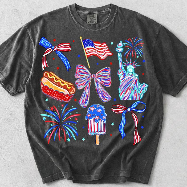 K&ouml;nnte beinhalten: Dunkelgraues T-Shirt mit patriotischen Illustrationen. Designs umfassen eine wehende amerikanische Flagge, die Freiheitsstatue, Feuerwerk, Schleifen, einen Hotdog und ein Eis am Stiel, alles in Rot, Wei&szlig; und Blau.