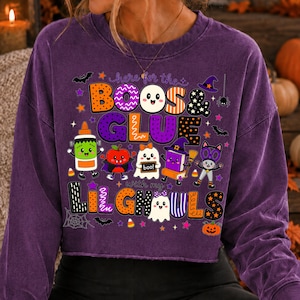 K&ouml;nnte beinhalten: Ein violettes Langarm-Sweatshirt mit Rundhalsausschnitt und einer bunten Halloween-Grafik mit dem Text "here for the BOOS & GLUE with my LIL GHOULS". Das Design zeigt verschiedene Zeichentrickfiguren wie Geister, Kleber und eine Katze.