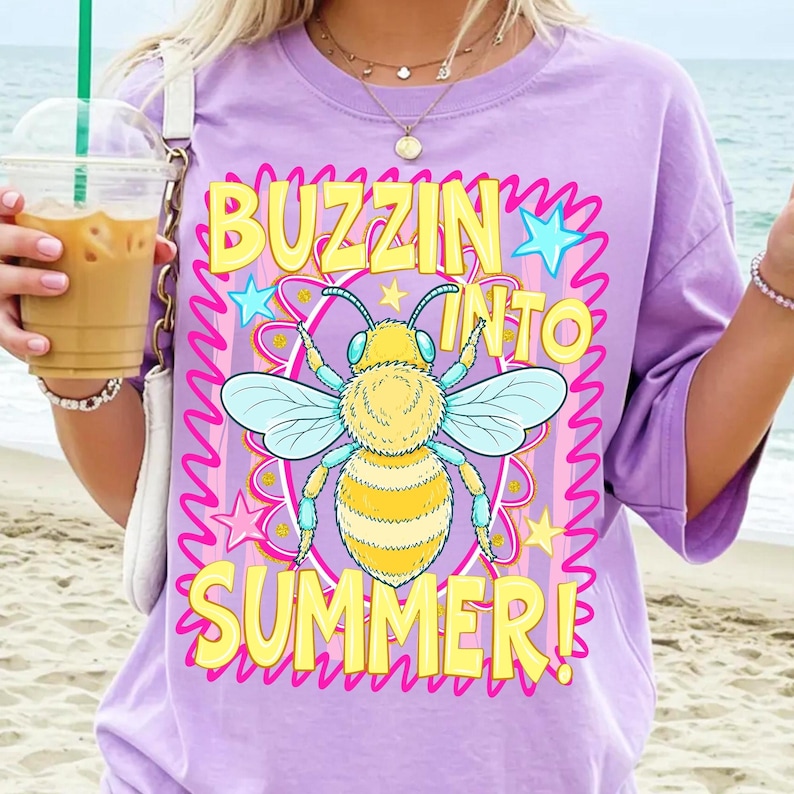 K&ouml;nnte beinhalten: Ein lila T-Shirt mit einer Biene und dem Text "Buzzin' Into Summer!" in Gelb. Die Biene ist gelb und blau, umgeben von rosa Sternen und einem wellenf&ouml;rmigen Rand. Das Shirt hat einen l&auml;ssigen, &uuml;bergro&szlig;en Stil.