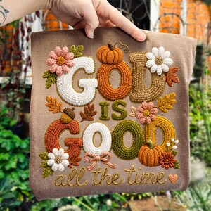 Pu&ograve; includere: Una t-shirt marrone con ricamo testurizzato che recita "GOD IS GOOD all the time" in filati bianchi, arancioni, verdi e gialli. Il design &egrave; decorato con zucche, margherite, ghiande e un piccolo cuore ricamati.
