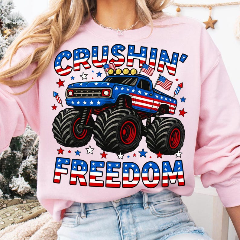 K&ouml;nnte beinhalten: Hellrosa Sweatshirt mit einem blauen Monstertruck-Design, das rote, wei&szlig;e und blaue patriotische Farben aufweist. Der Text "CRUSHIN' FREEDOM" wird in einer passenden patriotischen Schriftart mit Sternen und Feuerwerksk&ouml;rpern angezeigt.