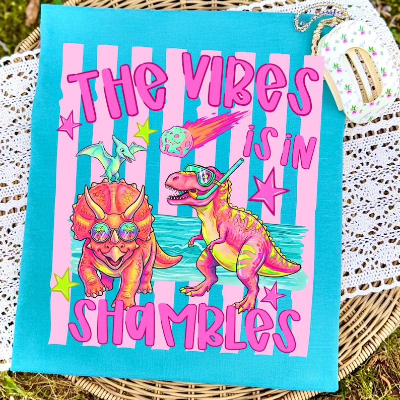Peut inclure: Un sac fourre-tout turquoise avec un motif &agrave; rayures roses et blanches repr&eacute;sentant des dinosaures de dessins anim&eacute;s et le texte "THE VIBES IS IN SHAMBLES" en rose. Le sac a une bordure en dentelle blanche.